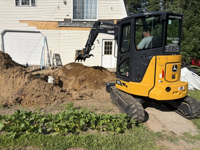 Nate's Septic branded John Deere 35 mini excavator digging beside a home's side entry on a deck support install in New Ipswich, NH